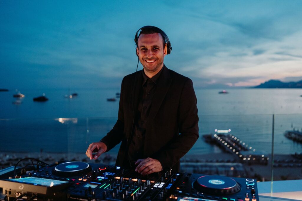 Brice Adel, wedding and event DJ based in Marseille, smiling on the decks on a rooftop overlooking the Bay of Cannes